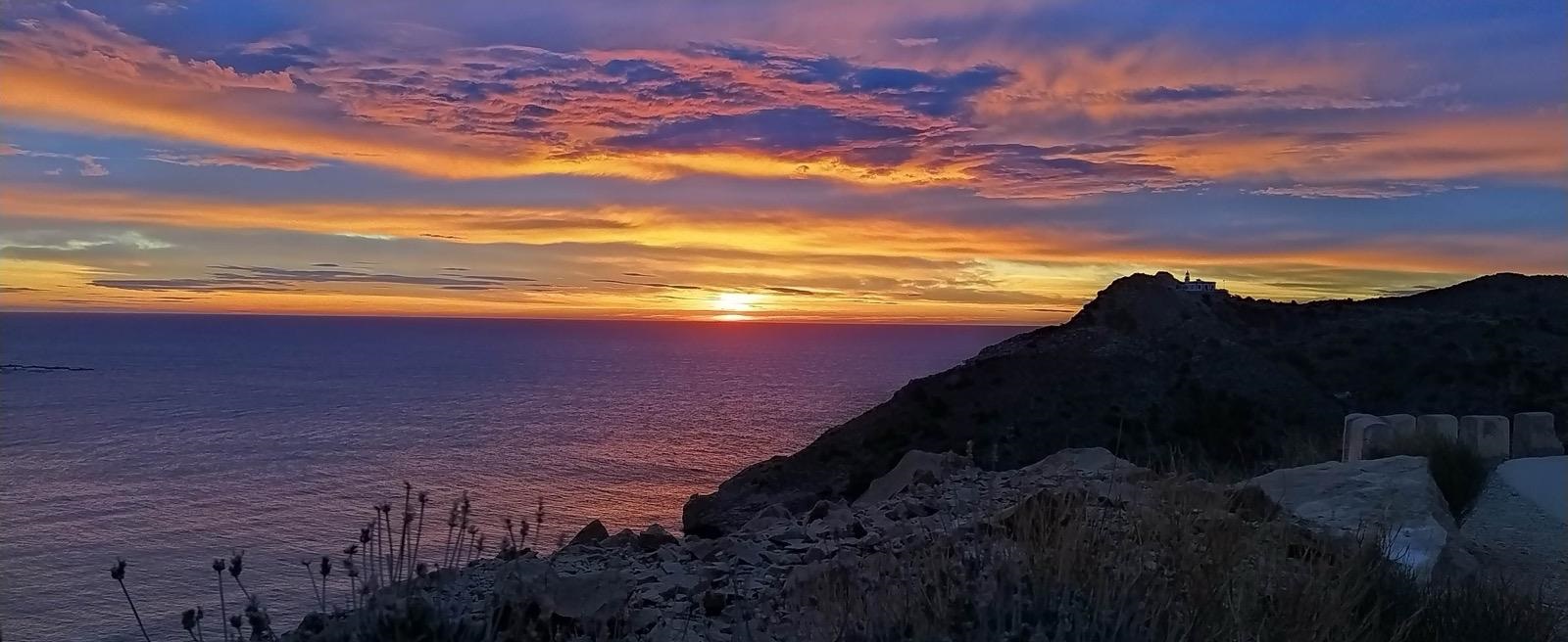 Amanecer sobre el faro del Albir con el cielo teñido de tonos naranjas, lilas y azules reflejándose sobre el Mediterráneo.