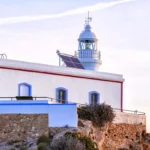 Faro del Albir sobre el acantilado al atardecer, con fachada blanca y detalles en azul y rojo.