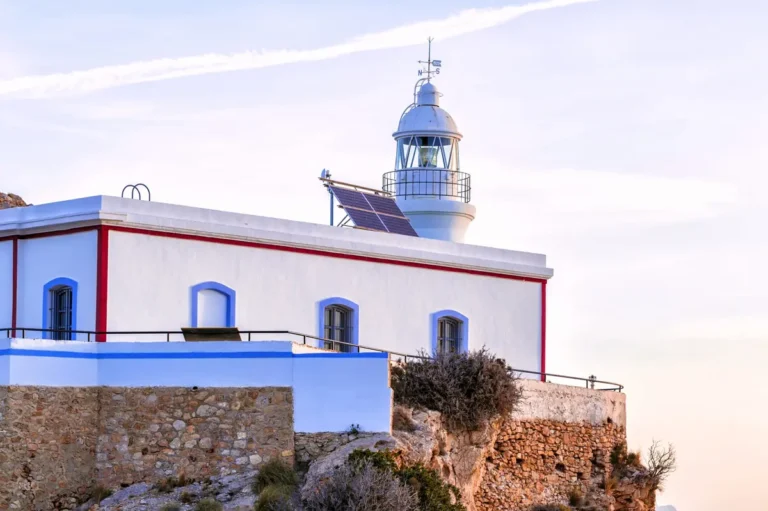 Faro del Albir sobre el acantilado al atardecer, con fachada blanca y detalles en azul y rojo.