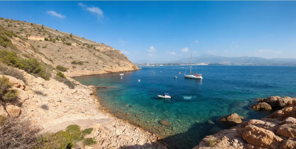 Cala de la Mina, en El Albir, con aguas turquesas y transparentes, veleros fondeados y los acantilados de la Serra Gelada al fondo.