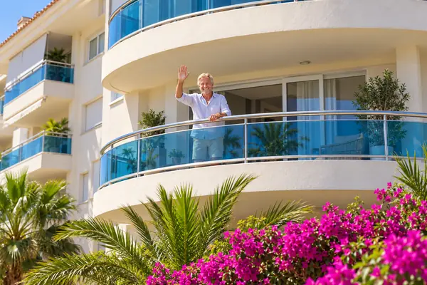 Pasar temporadas en El Albir: un apartamento para quedarse Hombre saludando desde el balcón de un apartamento, entre palmeras y buganvillas